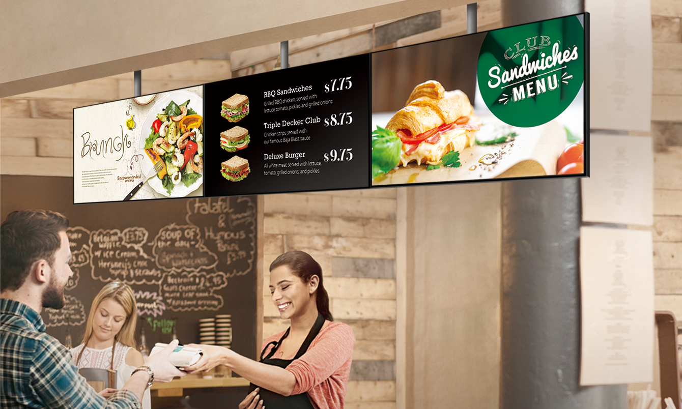 The staff in a sandwich store is handing a sandwich to a customer. The SM5J series showing a menu board is installed above them, displaying sandwich menus with brunch promotions.