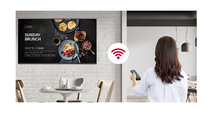 A woman is holding a remote controller and the display is connected in Wi-Fi. The woman is conveniently distributing content or update SW by using a remote controller since the display is connected in network thru Wi-Fi. 