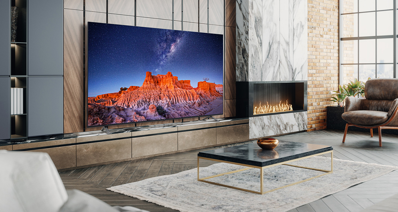 A large TV is placed on a living room wall decorated with a fireplace. Scenery of a mountain at night in which the Milky Way flows is bright and vivid on the TV screen.