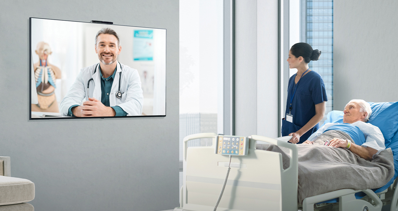 A patient is lying in the hospital room, and using the Smart Cam Pro on the wall, the doctor is teleconsulting by seeing the patient remotely.