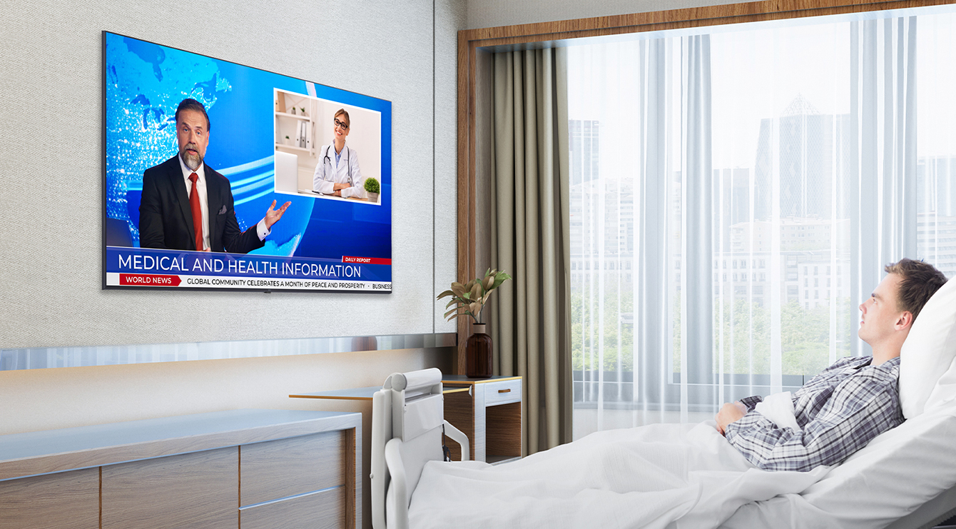 The patient is lying in the hospital room’s bed and watching the TV hanging on the wall.