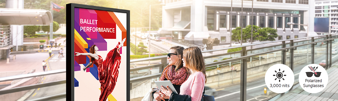 A large-sized display is installed on the street, and women are looking at a vivid-quality advertisement on the screen.