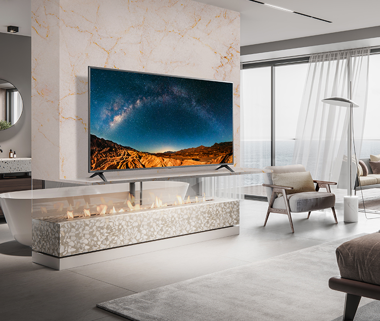 In a simple bedroom overlooking the sea, there is a TV on a wall shelf. The blue sea scenery appears bright and clear on the TV screen.