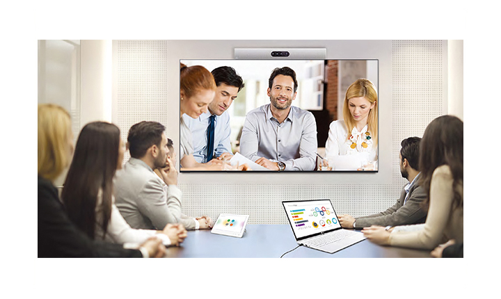 people are gathered in a conference room, having a virtual meeting with other people who are appearing on the screen.