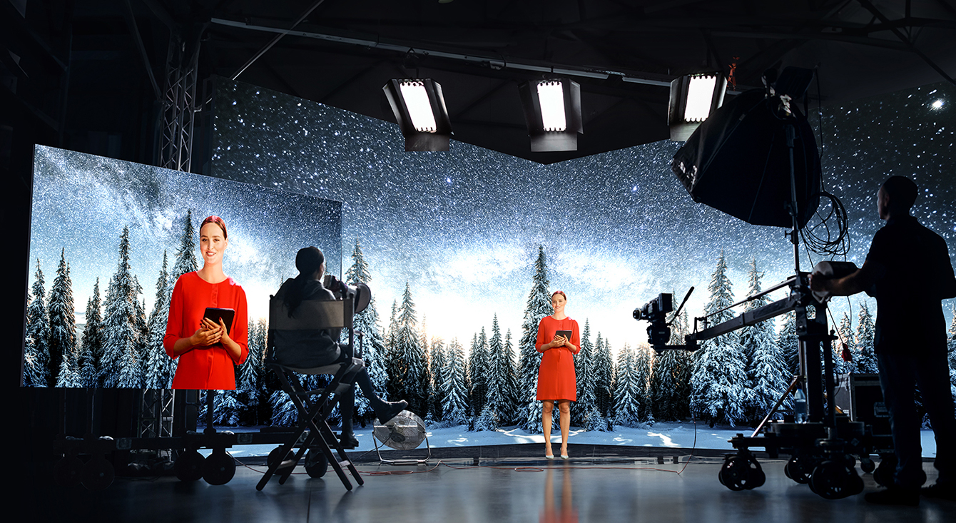 A woman is seen filming in front of a large cube-shaped LED screen. The LED displays a sparkling starlit snowy background, sharply contrasting with the woman's red dress, creating a vivid image on the large monitor.