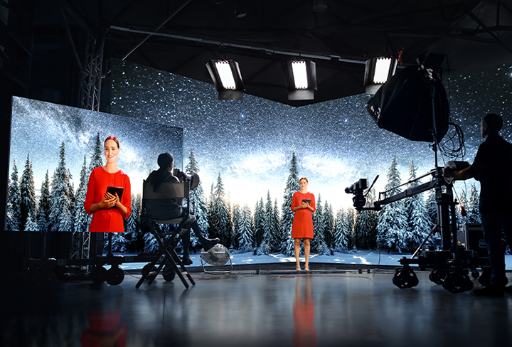 A woman is seen filming in front of a large cube-shaped LED screen. The LED displays a sparkling starlit snowy background, sharply contrasting with the woman's red dress, creating a vivid image on the large monitor.