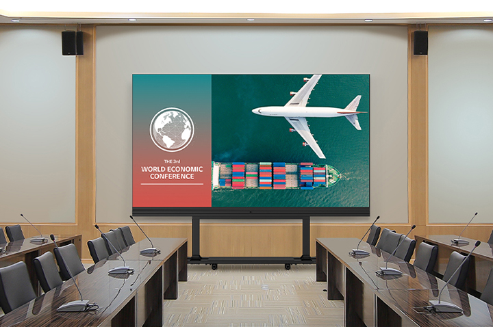In a spacious meeting room with long tables on both sides, a LABA series which is TAA (Trade Agreements Acts) compliant All-in-One LED model is placed against the wall. The LABA screen displays meeting content vividly. 