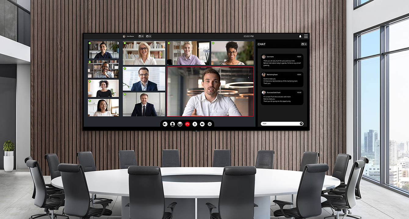 A wide LED screen is installed on the wall of a meeting room with a large round table. The expansive LED screen shows an ongoing video conference.