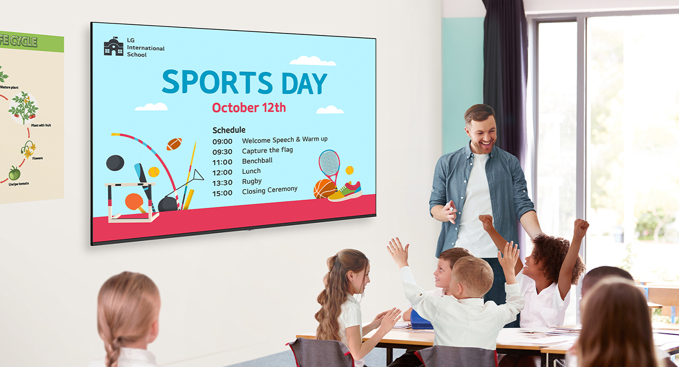 In a classroom, there are a teacher and students, and UM340E(TV Signage for school) is installed on the wall. The screen of the UM340E displays an event schedule, and the students are looking at it and enjoying themselves.