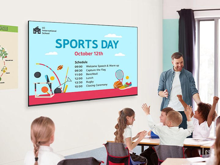 In a classroom, there are a teacher and students, and UM340E(TV Signage for school) is installed on the wall. The screen of the UM340E displays an event schedule, and the students are looking at it and enjoying themselves.