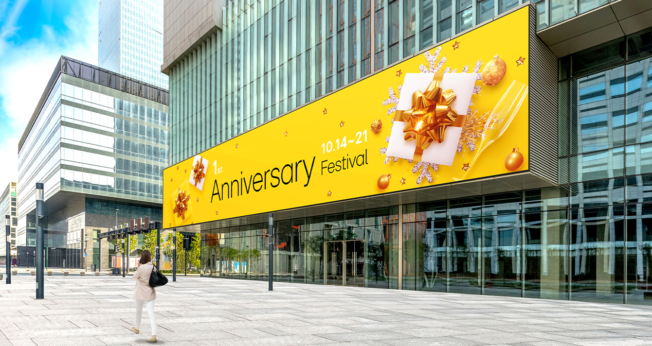 A large LED screen is installed on the exterior wall of the building entrance, and a woman is watching the advertisements on its screen. The screen remains clear and visible despite the bright sunlight.