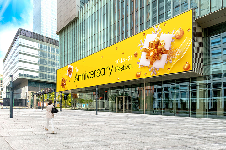 A large LED screen is installed on the exterior wall of the building entrance, and a woman is watching the advertisements on its screen. The screen remains clear and visible despite the bright sunlight.