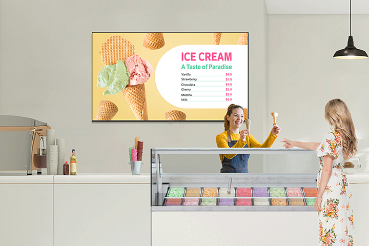In an ice cream shop, a UM340E is installed on the wall. The screen of the UM340E displays the menu prominently, and the customer is choosing her ice cream.