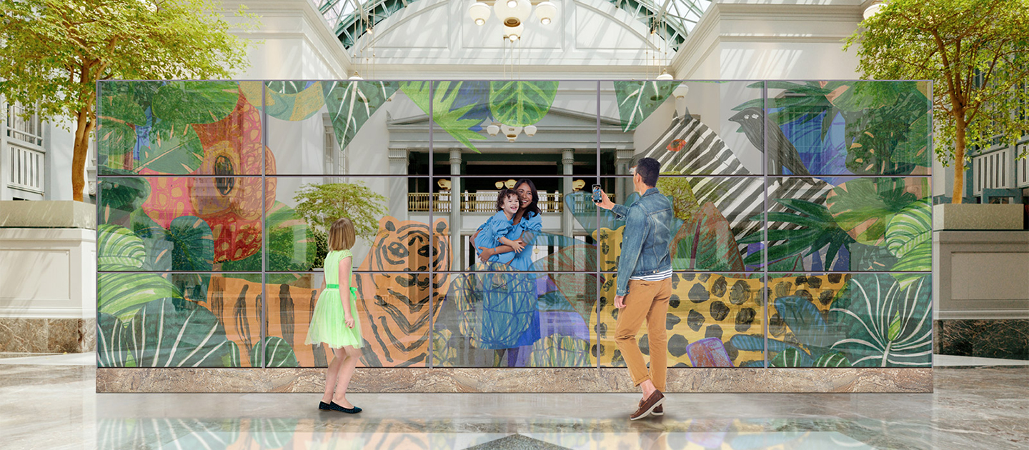 A family is taking a photo with a transparent OLED wall made up of 3ⅹ5 55EW5P-M indoors. The father, standing in front of the wall, is taking a picture of the mother and child, who are standing behind the wall with animals and plants naturally displayed on the screen.